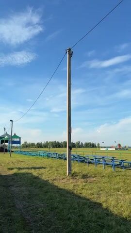 A drone is captured in flight against a bright blue sky with wispy white clouds. The drone is white and appears to be a remote-controlled aircraft. The drone is flying over a grassy field. In the foreground, a concrete utility pole with wires is visible. In the middle ground, rows of blue benches with green railings are set up in the field. In the background, a line of trees forms the horizon. The overall scene suggests an outdoor event or a recreational area where drone flying might be permitted.