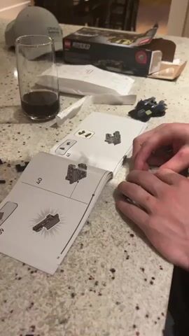 A person assembles a Lego Speed Champions kit on a speckled countertop. The instructions are open to pages 41 and 42. They use their hands to add a small Lego piece to a larger structure. A partial Lego structure sits to the side. Nearby, a glass with a dark liquid, a baseball cap, the Lego box, and packaging materials are visible.