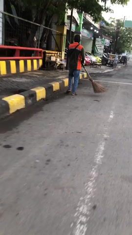 The video shows a street cleaner sweeping a dirty road. The cleaner is wearing an orange and green uniform. The road is paved with asphalt and has yellow and black painted curbs. There are several parked cars on the side of the road and some buildings in the background. The overall impression is one of urban cleanliness efforts.