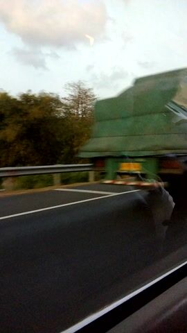 The video is a vertical, somewhat blurry, first-person perspective shot taken from inside a vehicle. The view is predominantly of a road with lane markings and what appears to be a construction site to the left, covered in green tarpaulin. A large vehicle, possibly a truck or trailer, is visible within the construction area. Trees with autumn foliage are also present. The sky is overcast. The video appears to be filmed from a moving vehicle, as indicated by the motion blur.