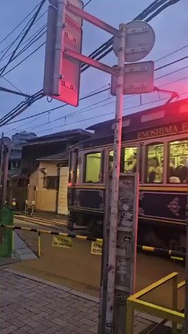 A train passes through a railroad crossing at dusk. The train is dark blue with gold trim and has lit windows, showing passengers inside. A red signal light is illuminated above the crossing, and yellow barriers are lowered. The crossing is paved with bricks and the sky is a dusky blue.