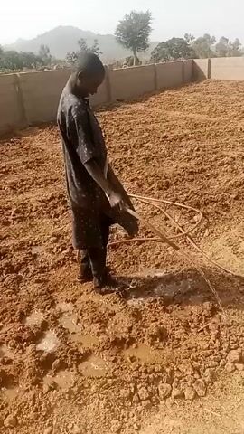 The video shows a person watering a large plot of plowed, muddy land. The person is wearing a dark, patterned shirt and pants, and sandals, standing in muddy areas as they water. They are holding a long, beige hose and directing the water onto the dry dirt. The plot is bordered by a short, tan-colored wall on one side, with trees and distant hills visible in the background under a hazy sky. The overall scene suggests agricultural work or land preparation in a rural environment.