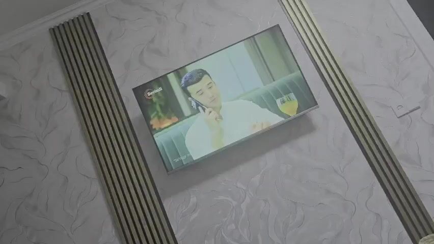 This media file captures a television screen displaying a scene in what appears to be a commercial or a show. The on-screen image features a young man with short dark hair, wearing a white collared shirt, seated on a green upholstered sofa. He is holding a smartphone to his ear with his left hand and looking slightly down and to his right. To his right, on the sofa, is a clear glass containing a yellow beverage, possibly juice or a cocktail. The background of the on-screen scene is softly blurred, suggesting a shallow depth of field, with hints of green foliage and a muted interior. A logo, possibly for a brand or channel, is visible in the top left corner of the screen, with the text "Sovuqli" in a circular emblem. Below the man, there is text that reads "Sotilayotgan".

The television itself is mounted on a wall with a patterned wallpaper. The wallpaper has a light, off-white base with a subtle, swirling, organic pattern in a slightly darker shade. Flanking the television are vertical decorative panels consisting of narrow, dark wooden slats with thin gold stripes in between. These panels extend from the top of the frame down towards the bottom. The overall setting suggests an indoor environment, likely a living room or a well-decorated space. The lighting appears to be artificial, consistent with an indoor setting at any time of day. The man on screen appears calm and engaged in his phone conversation. There are no visible weather cues or explicit location indicators beyond the on-screen text and the context of Uychi, Uzbekistan.