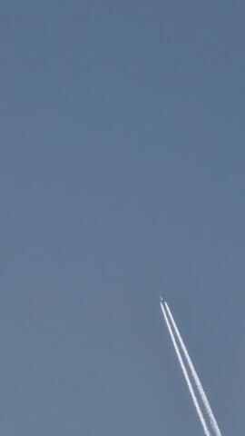 A commercial airplane is flying diagonally upwards across a clear blue sky, leaving two contrails behind it. The airplane is a tiny white speck at the top of the image, with the contrails stretching downwards and slightly to the right. The sky is a uniform light blue color with no clouds visible. The video is a static shot from a low angle, looking up at the airplane.