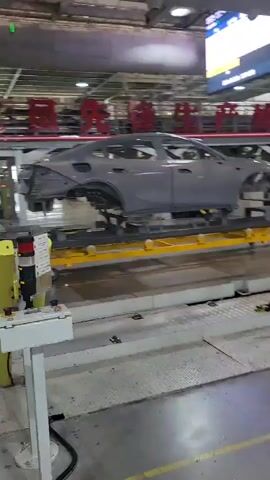 The video shows an automotive assembly line in Yufengshan, China. The primary subject is a grey car body, mid-assembly, moving along a conveyor system. It appears to be in the painting or pre-painting stage, as it's mostly bare metal with a primer coat. The car is mounted on a yellow, wheeled platform.

The setting is a large, industrial factory. Overhead, there are red Chinese characters displayed on a lighted sign, though they are not fully legible. Various pieces of factory equipment, tools, and parts are visible in the background, including robotic arms and hanging infrastructure. A yellow and grey control panel with a flashing red light is positioned in the foreground, suggesting an active workstation.

The activity depicted is the continuous movement of the car body through the manufacturing process. The lighting suggests an indoor, well-lit environment, typical of a factory operating at any time of day. There are no people clearly visible in the frame, reinforcing the impression of an automated or semi-automated production line. The overall mood is one of industrial efficiency and production.