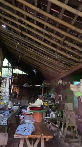 The video pans across the interior of what appears to be a rustic, open-air structure with a bamboo-slatted roof. The space is cluttered with various objects, suggesting it might be a kitchen or storage area. Visible items include cooking pots and utensils hanging on the walls, plastic containers, and what appears to be a pile of grain or sand in a corner. A wooden table is central to the scene, with some fabric and plastic items on it. The overall impression is one of a lived-in, possibly rural, environment.