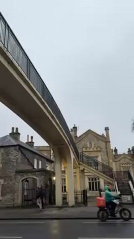 The video depicts a street scene on an overcast day. A concrete pedestrian overpass with black railings stretches across the road, connecting two buildings. One building is a traditional stone structure with gabled roofs and multiple chimneys. The other building features a set of stairs leading to an elevated walkway. A few people stand on the sidewalk near the stone building, talking. Cars pass on the wet road below the overpass. A red car moves from right to left, and a silver car follows.