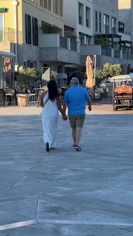 A couple walks hand-in-hand away from the camera across a paved outdoor area. The woman on the left wears a long, flowing white dress and has dark, long hair. The man on the right wears a blue t-shirt and khaki shorts. Behind them, buildings with stone and stucco facades line the area, with outdoor seating and umbrellas visible. Other people are seen in the background, and a small orange vehicle is parked on the right side of the frame.