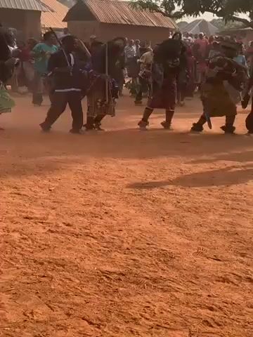 The video shows a group of people in what appears to be a traditional performance or ceremony in a rural setting. The performers are dancing on a reddish-brown dirt ground, with a crowd of people watching in the background. The dancers are wearing traditional clothing, some with masks or headdresses, and seem to be moving in a coordinated fashion. The background includes simple buildings with thatched roofs, suggesting a village environment. The day appears bright, with shadows indicating the position of the sun.