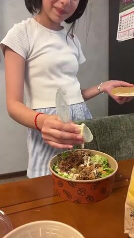A young girl with dark hair, wearing a white t-shirt and blue and white striped shorts, stands at a wooden table. She is holding a small plastic container with a clear lid in her left hand, from which she is pouring something into a salad bowl. The salad bowl, a disposable paper container with an orange and brown pattern, is filled with salad. In her right hand, the girl holds a piece of flatbread or a cracker. To the right of the salad bowl, there is a brown paper grocery bag with an image of the Colosseum. In the foreground, a clear plastic lid sits on the table. Behind the girl, a dark refrigerator is visible, with a calendar hanging on it. The environment appears to be a kitchen or dining area.