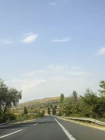 A car is driving on a highway. The highway has two lanes and a median. The median is lined with trees and a guardrail. The car is driving through a rural area with hills and trees. The sky is blue with some clouds.