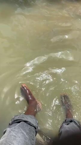 The video shows a person's legs and feet submerged in murky water. The person is wearing gray pants. The water is a light brown color and appears to be shallow, as the person's ankles and feet are visible. Sunlight reflects off the surface of the water, creating bright spots. The person's feet are moving slightly in the water, causing ripples. The background is out of focus and appears to be more murky water or possibly the edge of a body of water.