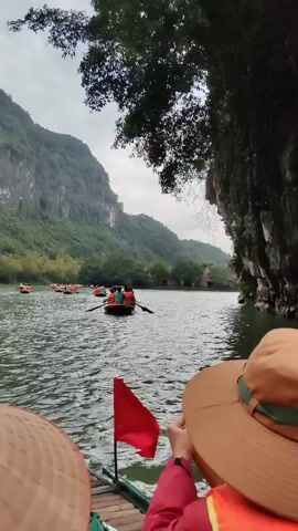 The video shows a first-person perspective from a boat on a river, with numerous other boats visible ahead. The boats are filled with people wearing life jackets, and they are rowing through the water. Lush green mountains rise in the background under a cloudy sky. Vegetation, including trees, is visible on the river banks. The foreground includes portions of two hats, one a traditional Asian conical hat and the other a Western-style hat, suggesting the viewer is in one of the boats. A small red flag is also visible in the foreground, possibly attached to the boat.