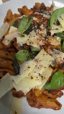 A close-up shot focuses on a plate of pasta, likely penne, covered in a rich, reddish-brown sauce. Mixed within the pasta are pieces of sautéed mushrooms. The dish is generously garnished with large, thin flakes of Parmesan cheese, fresh green basil leaves, and a sprinkle of red pepper flakes. The lighting is bright, highlighting the textures and vibrant colors of the ingredients. No people are visible, and the setting appears to be a dining surface, possibly a table.