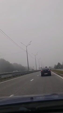 The video captures a view from inside a vehicle traveling on a highway on a foggy day. The road is paved with multiple lanes, and a guardrail is visible on the left side. Several streetlights line the highway, and trees shrouded in fog can be seen on the left. A dark-colored sedan is driving ahead in the right lane, and other vehicles are faintly visible in the distance. Traffic lights and speed limit signs (50) are also present along the road. The overall atmosphere is hazy and overcast, suggesting low visibility.