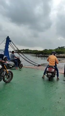 This video shows a ferry with multiple people and motorcycles on it. In the foreground, a man on a red scooter wearing a mask and helmet is facing away from the camera, looking out at the water. To his left, another man on a scooter is also looking out at the water. In the background, there is a body of water with trees on the shore. The sky is cloudy and overcast. The ferry appears to be in motion, possibly docking or departing.