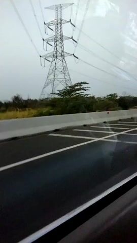 A tall, lattice-style electrical tower stands in the background under a cloudy sky. Power lines extend from the top of the tower. In the middle ground, a line of trees and dry vegetation is visible. A concrete barrier separates the foreground from the middle ground. The foreground is an asphalt road with white lane markings. The road appears wet, suggesting recent rain. The perspective suggests the video was captured from a moving vehicle, likely looking out a window. No people or animals are visible.