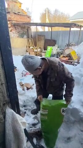 The video shows a man kneeling in a snowy, cluttered yard. He is wearing a gray beanie, a camouflage jacket, and dark boots.  He is holding a large green bag with the word "kitti" printed on it and appears to be rummaging in the snow. The area around him is covered in snow and scattered with various objects including boxes, a green container, and trash. In the background, there's a partially destroyed brick structure, a wooden frame, and a corrugated metal fence. The weather appears to be cold and overcast.