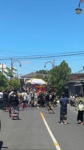 A group of people walking down a street on a bright day is shown in this video. They are carrying what appears to be a small, ornate structure, possibly a shrine or a float, decorated with white and gold. The individuals are dressed in a mix of traditional and casual clothing, with many wearing sarongs. The street is lined with buildings and trees, and there are street lamps with curved designs. Overhead, power lines crisscross the clear blue sky. One man at the edge of the crowd carries a framed picture. The scene suggests a procession or ceremony.