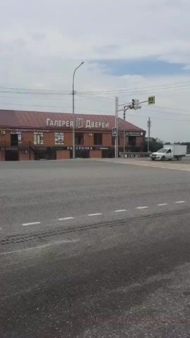 The video shows a wide, empty intersection with asphalt pavement and painted lane markings. In the background, a two-story brick building with a red roof has large text on its facade that reads "ГАЛЕРЕЯ ДВЕРЕЙ" (Gallery of Doors). A white pickup truck with a white cargo box is visible on the road. A white sedan is also seen driving through the intersection. Traffic lights and pedestrian crossing signs are present. The sky is overcast with clouds.