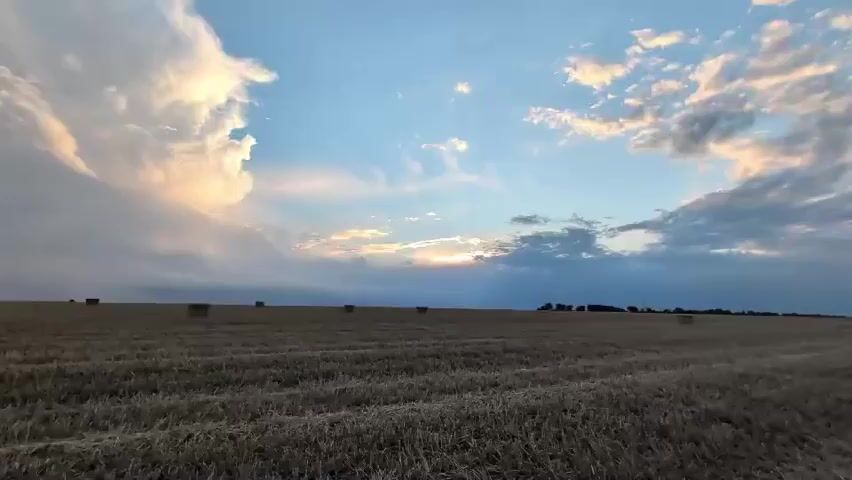 The video shows a drone's-eye view of a harvested field under a cloudy sky with the sun breaking through. The field is dry and consists of stubble with scattered round bales of hay. The drone moves forward, giving a sense of depth and distance. The sky is a mix of blue and white clouds, with some tinged with orange and pink from the setting or rising sun. Trees and a faint structure are visible in the distance.