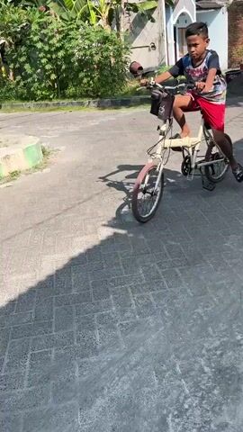 A young boy rides a cream-colored folding bicycle on a paved street. He wears a dark blue t-shirt with multi-colored graphics, red shorts, and black sandals. The bicycle has a basket attached to the handlebars. Behind him, a black scooter with two passengers, a person driving and another person behind, turns the corner. The driver wears a white hoodie and a light green headscarf, while the passenger wears a dark hoodie and a black backpack. The surroundings consist of houses, greenery, and trees. The weather appears sunny.