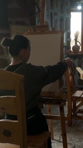 The video shows the interior of a room with stone walls, possibly an art studio or workshop. A young woman is seated on a wooden chair in front of an easel, sketching on a canvas. The room is illuminated by natural light coming through two windows, as well as several light bulbs hanging from the ceiling. Other wooden furniture is scattered throughout the room, including a table, chairs, and stools. On one of the tables, there's a decorative object resembling a relief of a leaf. The overall impression is one of a rustic, creative space.