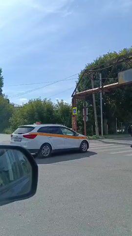 A white station wagon with an orange stripe drives over a pedestrian crossing. A dark-colored station wagon is behind it. A person is standing on the side of the road. The sun is shining.