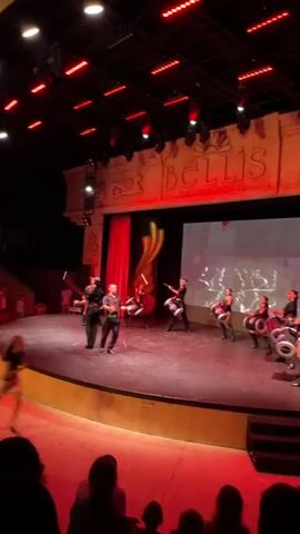 A group of performers is on a stage under red lighting. A man in the center is juggling batons, while several other performers in black outfits are playing large drums. The stage has a red curtain to the left and a large screen with a light show behind the performers. The words "BELLIS DELUXE HOTEL" are visible on a wall above the stage. A child runs across the lower-left foreground of the image, partially obscuring the audience seated in the theater.