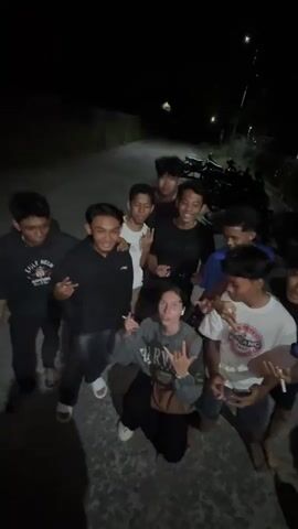 A group of eight young men and one young woman are gathered outdoors at night. They appear to be posing for a photo and are making hand gestures. The individuals are dressed casually. The environment is dark, suggesting it is nighttime. There is a road or path behind them, and some vegetation is visible. A motorcycle is parked in the background. The overall mood seems energetic and celebratory.