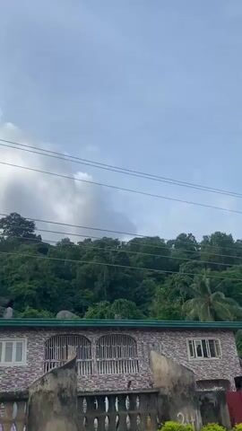 The video shows a vehicle driving down a road in front of a heavily forested hillside. Several buildings, including a small, two-story house with a red gate, are visible on the right side of the road. There are a few palm trees and other green vegetation in the foreground and on the hillside. Power lines run across the sky, which is partly cloudy. The video appears to have been filmed on a bright, sunny day.