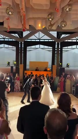 The video captures a wedding reception where a bride and groom are engaged in their first dance on a brightly lit dance floor. The bride is wearing a long white dress and the groom is in a dark suit. They are holding hands and facing each other, surrounded by wedding guests who are standing around the dance floor watching and taking pictures. In the background, there is a DJ booth with personnel visible behind it. The venue has a high ceiling with decorative disco balls and draped fabric. The room is framed by large windows offering a glimpse of the outside. The camera angle is from within the crowd of guests.