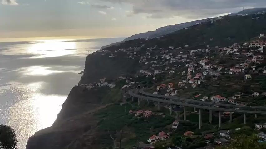 The video pans across a coastal hillside covered in houses with red-tiled roofs. A multi-level highway viaduct dominates the middle ground, with cars visible on its surface. The sea is on the left, reflecting sunlight. The sky is partly cloudy. The terrain is steep and green, with some terraced farming visible among the houses. The houses are clustered together, following the contours of the hill. The highway has many support pillars and curves around the hillside. The camera movement is a slow pan from left to right.