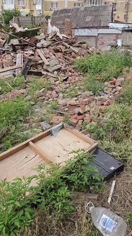 A pile of rubble is visible in the center of the image. The rubble consists of bricks, concrete, and wood. There is also some debris scattered around the rubble. In the foreground, there is a large piece of plywood with wooden framing. To the right of the plywood, a black object that appears to be a television is visible. A plastic water bottle is also in the foreground. In the background, there are buildings and trees. The overall scene suggests an area that has undergone demolition or some form of destruction.