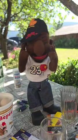 A puppet is shown outdoors. It is wearing a black baseball cap, a white t-shirt with the number 23 and a basketball design, and blue jeans. The puppet is standing at a table with several objects, including batteries, a glass, and what appears to be a container of food. The puppet is moving its hands in front of its face, and its mouth is open as if speaking or singing. The background shows trees and greenery, suggesting a sunny day.