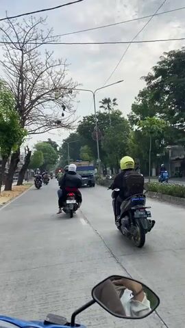 The video shows a street scene with several motorcycles and a truck. The perspective appears to be from another motorcycle, looking forward. In the foreground, the back of a motorcycle and its rider, wearing a white helmet, are visible. To the right, another motorcycle with a rider in a yellow helmet is also moving forward. In the middle ground, a truck is driving ahead, and several other motorcycles are scattered on the road. The environment appears to be urban or suburban, with trees lining the street and some buildings visible on the left. The sky is overcast. The video captures a moment of traffic flow on a road.