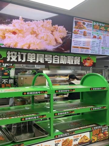 The video shows the interior of a fast-food restaurant. There are two staff members behind the counter, preparing food. Above them is a large digital menu board with images and prices of various food items. In front of the counter is a green metal shelf unit for order pickup. A person wearing a black shirt and baseball cap is standing in front of the counter, taking a photo of the menu with their phone. The color scheme of the restaurant is primarily green and white.