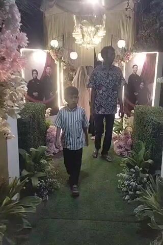 The video captures an outdoor event, possibly a wedding reception, illuminated by decorative lighting and a chandelier. Two large framed portraits of a couple in formal attire are prominently displayed on the left and right sides of a pathway covered with artificial grass. Lush floral arrangements, including pink and white flowers, are placed along the pathway and surrounding hedges.

In the foreground, a young boy wearing a striped shirt and black pants walks along the pathway. Behind him, a man in a patterned shirt and black pants follows. Further down the path, several people, including women in modest attire and headscarves, are visible, moving towards the background where the main event appears to be taking place. The ambiance suggests a celebratory and elegantly decorated setting.