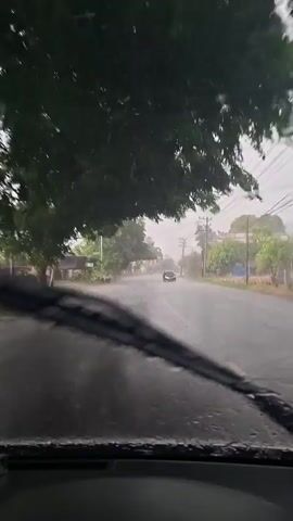 The video shows a view from inside a car driving on a wet road during a downpour. Raindrops are visible on the windshield, and the windshield wipers are intermittently sweeping the water away. Trees and buildings line the road, and a few other vehicles are seen in the distance. The sky is overcast and gray. The road appears to be paved with asphalt and has a yellow line marking the center. The overall impression is that of a rainy day drive through a rural or suburban area.