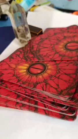 The video shows a close-up of a stack of tarot cards with a red, eye-like design on the back. A hand is seen lifting the top card, revealing the Nine of Wands from the Rider-Waite Tarot deck. The card depicts a woman standing with nine flaming wands. The background is slightly out of focus and appears to be a desk with various items, including a crystal. The lighting is soft and natural.