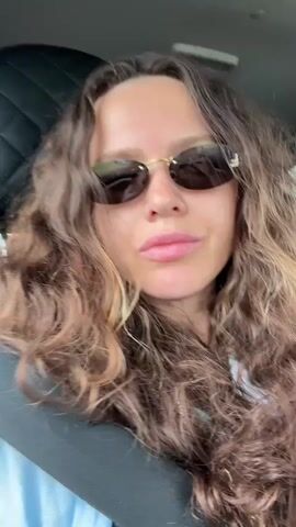 A young woman with long, curly brown hair and wearing sunglasses is in the passenger seat of a car. She is looking at the camera and smiling. She is wearing a light blue shirt and a seatbelt. Her left hand is resting on her chin and her right hand is resting on her chest. She is wearing several bracelets on her left wrist and a ring on her left index finger.