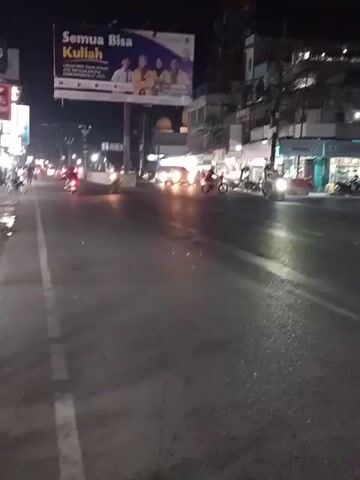 This media file captures a nighttime scene on a busy street in Bireuen, Indonesia. The camera is positioned low to the ground, likely from a vehicle, looking forward down a wide, paved road. The time of day is clearly night, with the sky being completely dark and artificial lights illuminating the scene. The weather appears to be clear, as there is no visible rain or fog affecting visibility.

The street is filled with activity. Numerous motorcycles, typical of Indonesian transportation, are visible in motion, their headlights and taillights creating streaks of light. Several cars are also present, including a silver hatchback that is particularly noticeable as it drives past, its brake lights glowing red. The road surface is wet in places, reflecting the streetlights and headlights, suggesting recent rain or ongoing street cleaning.

On the right side of the road, there are several buildings lining the street, appearing to be shops or commercial establishments. These buildings are illuminated with various lights, some with signage visible. One prominent billboard is suspended over the road on the left side, displaying an advertisement. The text on the billboard is in Indonesian and reads "Semua Bisa Kuliah" which translates to "Everyone Can Go to College." Below this main text are images of four people in graduation attire, implying the advertisement is for an educational institution. Other businesses have signs like "AROMA" visible.

The overall atmosphere is one of urban night life, with the constant movement of vehicles and the bright lights of businesses and advertisements. The presence of many motorcycles and the general streetscape suggest a typical busy road in an Indonesian city. No specific emotions are overtly conveyed, but the scene depicts a functional, active urban environment at night.
