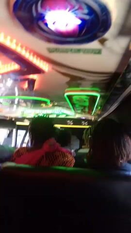 The video shows the interior of a vehicle, possibly a bus or van. The ceiling is decorated with colorful lights, including red and green LED strips, and a circular light with a spider design. Several passengers are seated, with their backs to the camera. One passenger in the foreground wears a pink headscarf and a patterned shirt. Another passenger on the right has dark hair. In the background, a man in a white shirt appears to be interacting with someone outside the vehicle, visible through a window. The lighting is dim, with the bright interior lights creating a vibrant atmosphere.