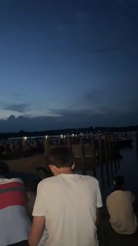 This is a vertical video of a group of young people sitting on benches outdoors at dusk. The sky is a deep blue with some scattered clouds. In the background, there is a body of water, possibly a lake or bay, with a wooden pier extending into it. Several boats are docked at the pier, and there are lights visible along the shore. A group of people are gathered on the pier and along the shoreline. The foreground shows the backs of three young people, two of whom are looking down at a phone. One person in the foreground is wearing a white t-shirt, another is wearing a dark t-shirt with a Patagonia logo, and the third is wearing a shirt with red and white stripes. The overall atmosphere appears to be relaxed and social, possibly at an outdoor event or gathering.