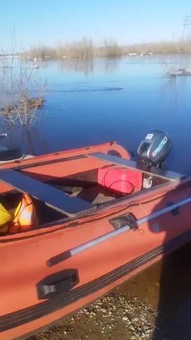 The video shows a flooded area on a sunny day.  The water reflects the clear blue sky. Patches of ice and snow are visible amongst the trees and bushes standing in the water. In the foreground, an orange inflatable boat with a motor is partially visible, resting on the muddy bank.  The water appears calm, with reflections of the surrounding vegetation. An electrical tower is seen in the distance.