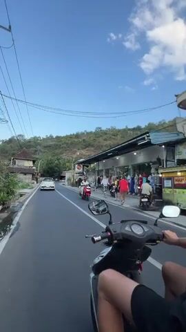The video is shot from the perspective of someone riding on the back of a motorcycle. The rider's legs are visible in the foreground. The motorcycle is traveling on a paved road. On the right side of the road, there are several shops with clothing racks outside. On the left side of the road, there are houses and some greenery. In the distance, there is a hill covered in trees. The sky is blue with some clouds. Other motorcycles and a white car are also visible on the road. The rider on the back of the motorcycle extends their right hand and waves it in the air.