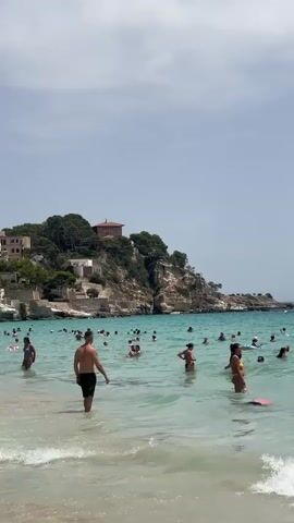 A wide shot captures a crowded beach scene under a bright, cloudy sky. The turquoise water is populated with numerous people swimming, wading, and floating. In the foreground, gentle waves wash onto a sandy shore. A man in dark shorts stands facing away from the camera, partially submerged in the water. To his left, closer to the shore and in focus, a man with short hair and a beard looks to his left, his shoulders and upper chest visible. In the background, a rocky, tree-covered cliff rises, topped with several buildings, one with a reddish-brown roof. The water extends to the horizon, with a few small boats visible in the distance. The overall impression is a lively and popular beach destination on a summer day.