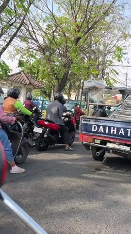The video captures a street scene with moderate traffic, likely in a Southeast Asian country given the prevalence of motorcycles. Several motorcycles are visible, with riders wearing helmets, some in casual attire. One motorcycle in the foreground has a rider in a bright green vest and another individual behind them. Another motorcycle, prominently featured in the center, is red and black. A white pickup truck with the brand name "DAIHATSU" is parked on the right side of the frame, partially obscuring other vehicles. The truck has a metal rack on its bed, and a light-colored tarp is draped over something in the truck bed. In the background, there are trees with lush green foliage, suggesting it is daytime. A building with a tiled roof and a small fence is also visible behind the traffic. The overall impression is of a typical urban street environment with active, albeit slow-moving, traffic.