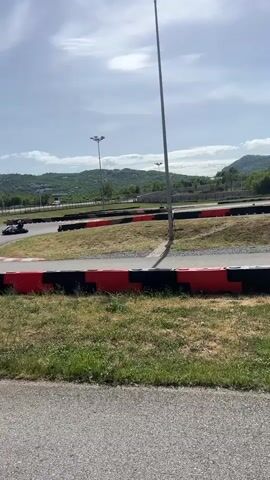 The video shows a go-kart track on a sunny day with mountains in the background. A person is driving a black and teal go-kart on the track, which is lined with red and black barriers. The camera is positioned behind a metal fence, partially obscuring the view. The track has several turns, and the go-kart is seen navigating one of them. The surrounding area consists of grass, light poles, and trees.