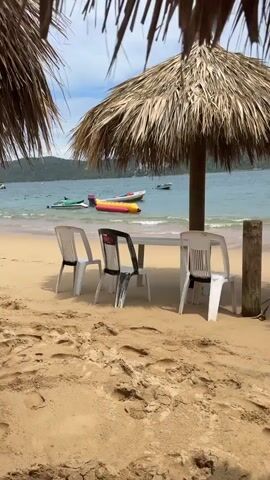 The video showcases a serene beach setting with several white plastic chairs and a table beneath a thatched umbrella. The sandy beach extends towards the ocean, where gentle waves lap at the shore. Several boats are visible in the calm blue water, including a yellow and red inflatable banana boat. The water is clear, and the sky is bright with some clouds. Footprints are visible in the sand, indicating recent activity. The scene suggests a tranquil day at the beach, possibly at a resort or a public beach area. The video slowly pans from left to right across the beach.